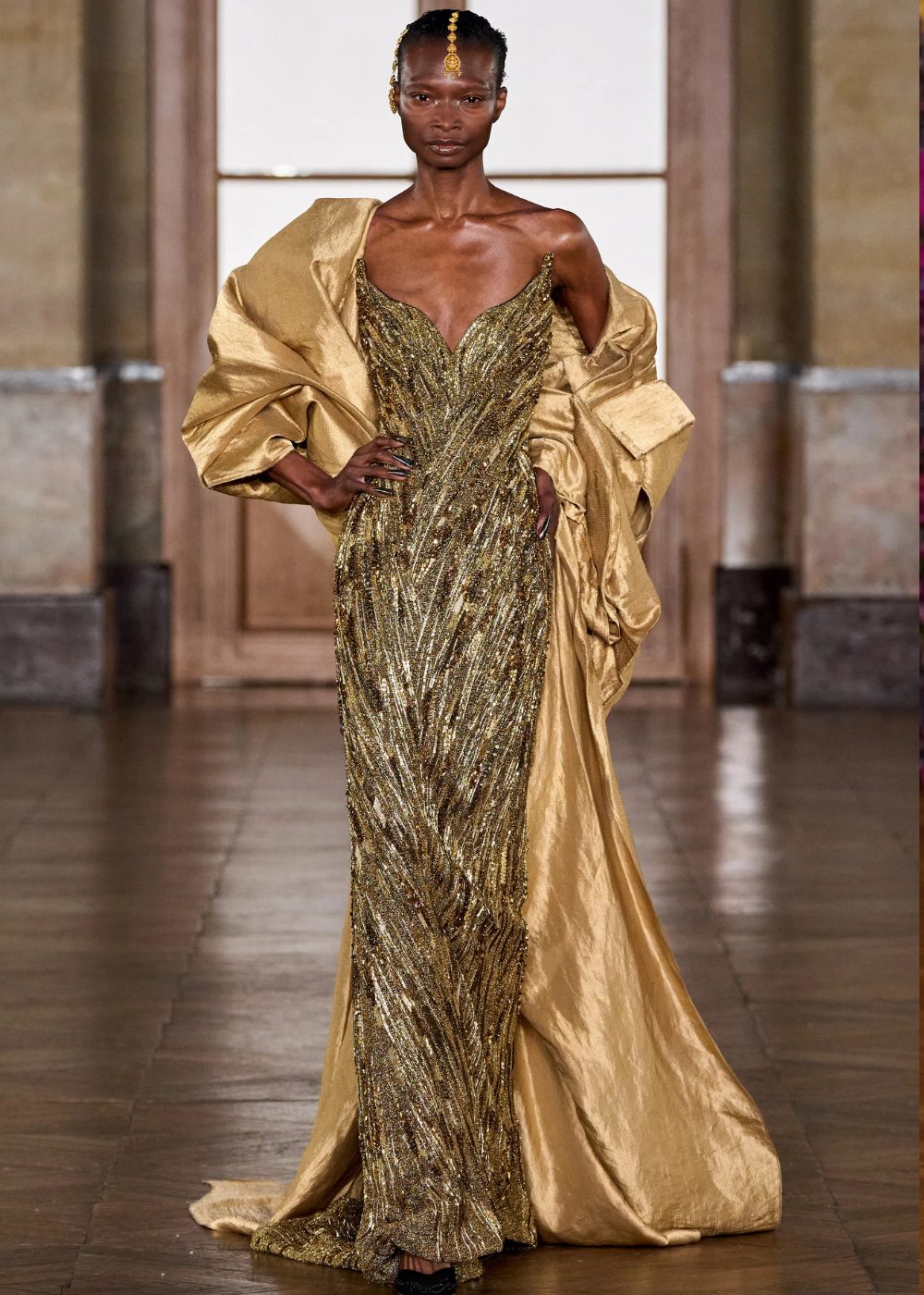 Modelo posa com vestido longo dourado bordado em linhas diagonais e cauda, complementado por capa de tecido acetinado,