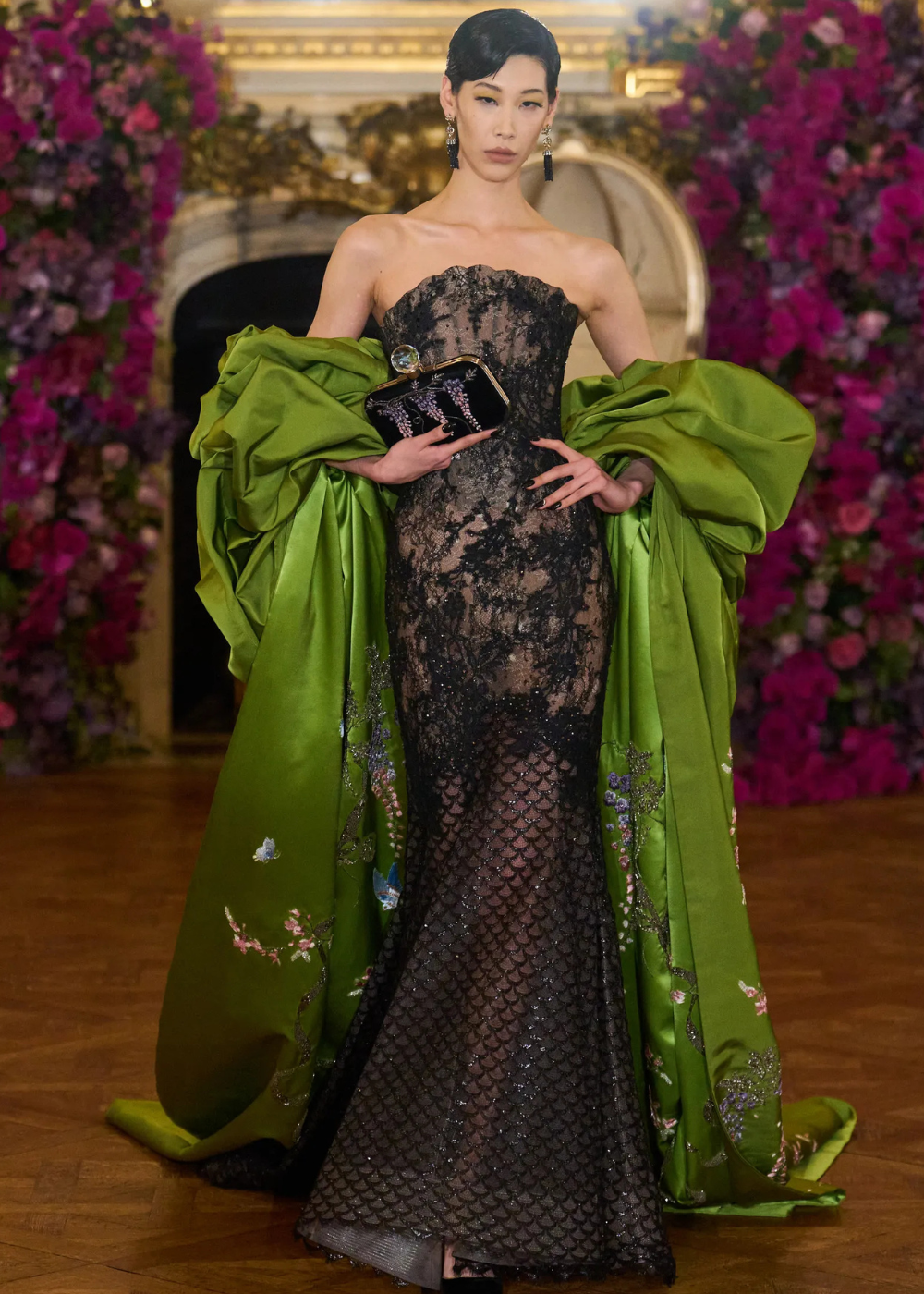 Modelo posa com vestido de alta-costura primavera 2026 em renda preta translúcida, cauda verde vibrante bordada e clutch