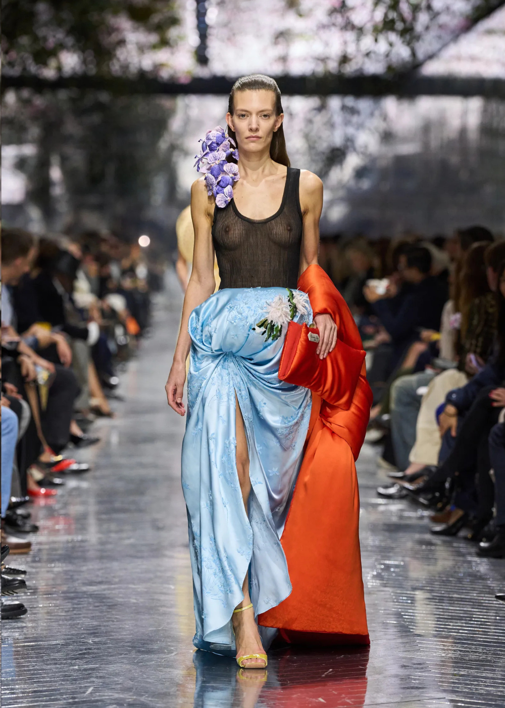 Modelo caminha com postura confiante usando body preto transparente, saia azul claro drapeada com fenda e maxi casaco