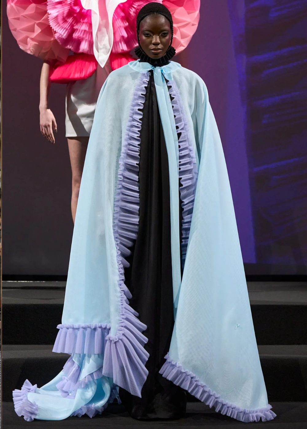 Modelo posa com capa fluida azul claro de tule e babados lilás sobre vestido preto, destacando tendências da temporada de