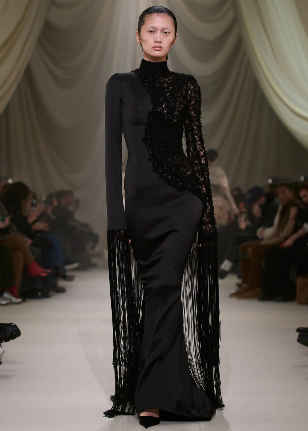 Modelo desfila vestido longo preto com mangas assimétricas, renda e franjas, evidenciando elegância e sofisticação nas