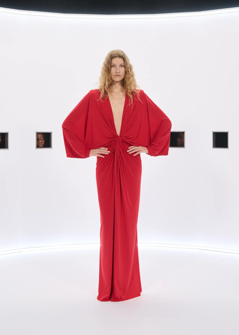 Modelo posa com vestido longo vermelho de tecido fluido, mangas amplas e drapeado central, evidenciando tendências da