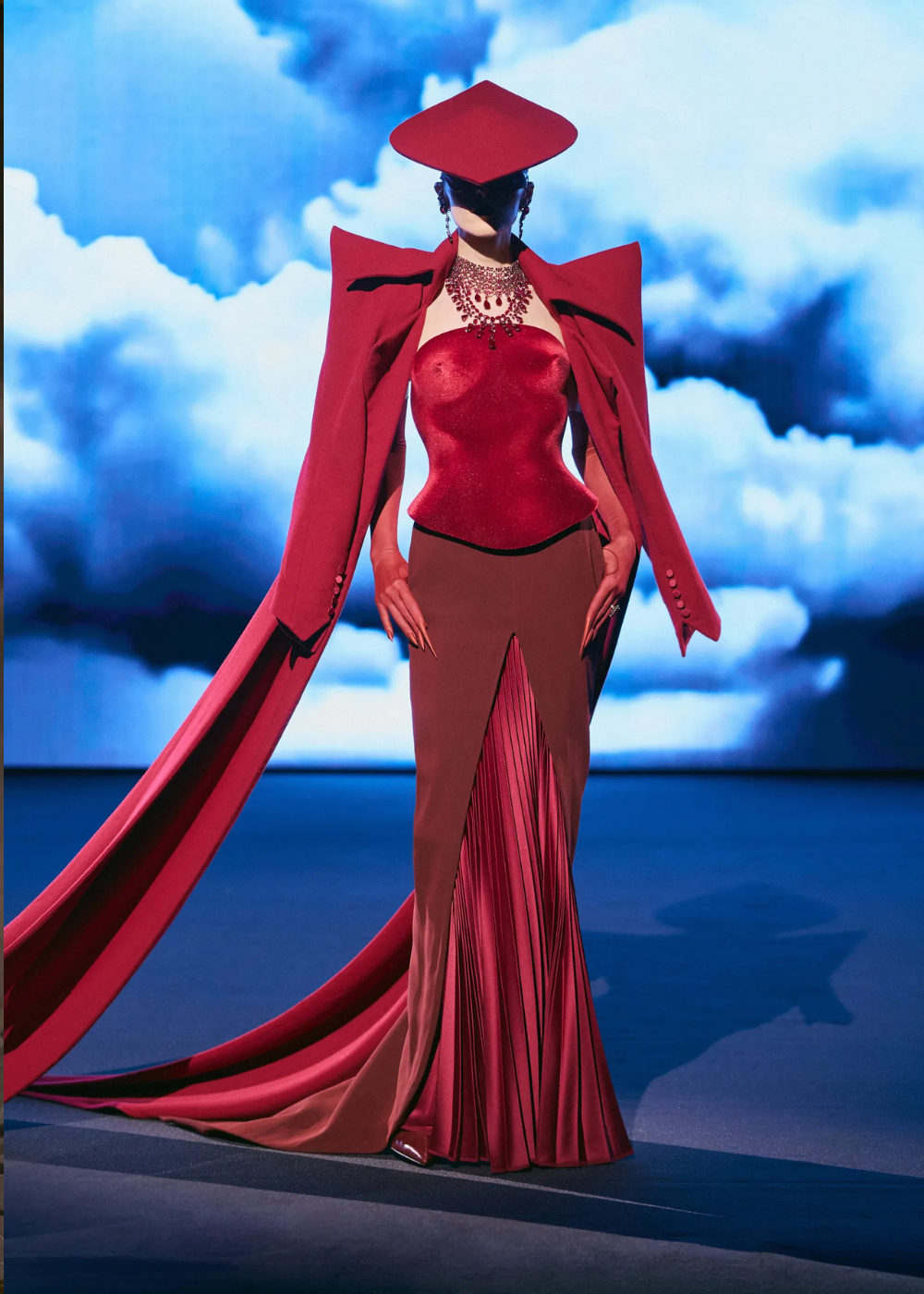 Modelo posa com look vermelho vibrante da alta-costura primavera 2026, combinando corset estruturado, saia plissada e capa