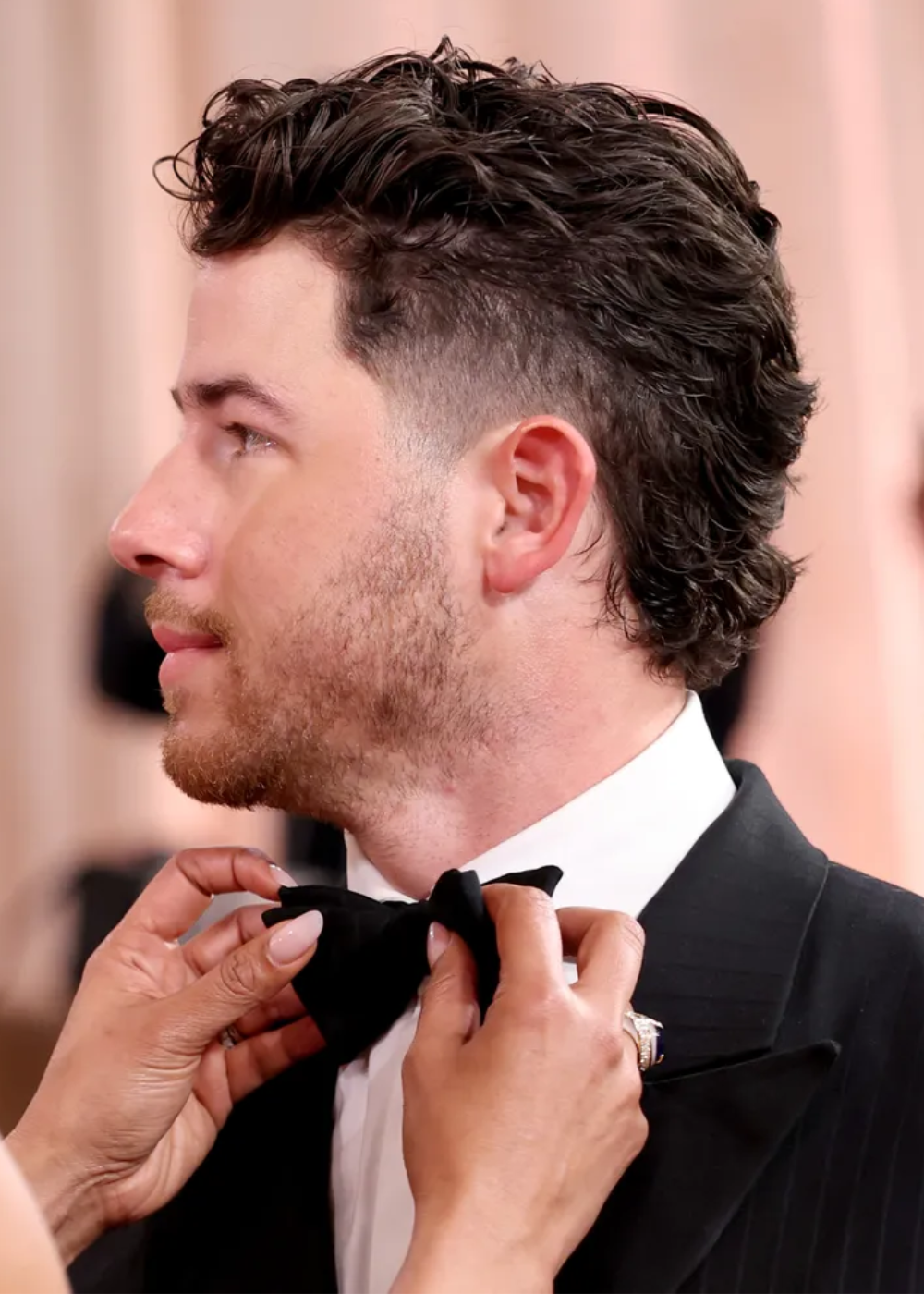 Homem com cabelo ondulado penteado para trás usa smoking preto de tecido acetinado e camisa branca.