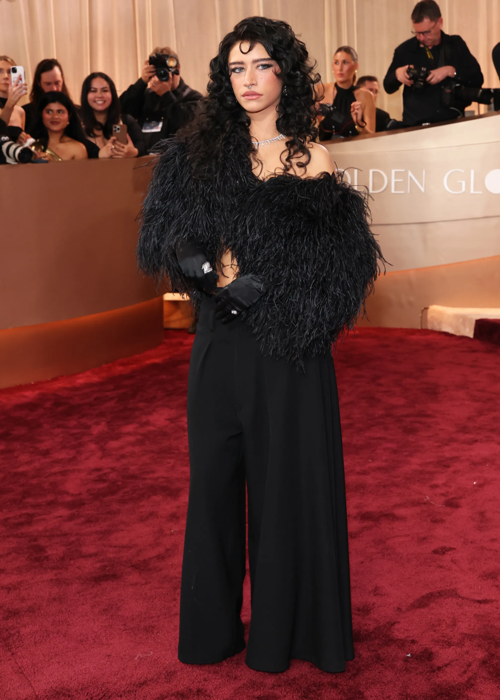 No Globo de Ouro 2026, modelo posa com look preto sofisticado, casaco de plumas volumoso, calça ampla e luvas elegantes.