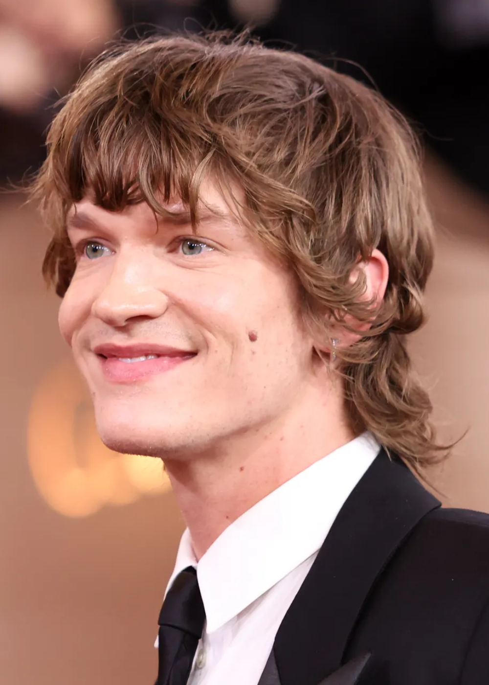 Ator sorri levemente com corte mullet e terno preto clássico de lapela em cetim, camisa branca e gravata.