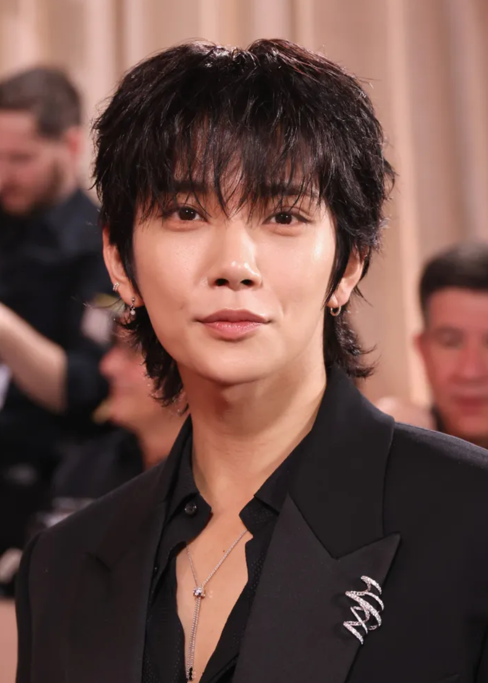 Homem posa para foto com blazer preto de modelagem clássica e broche prateado em espiral, estilo elegante e moderno.