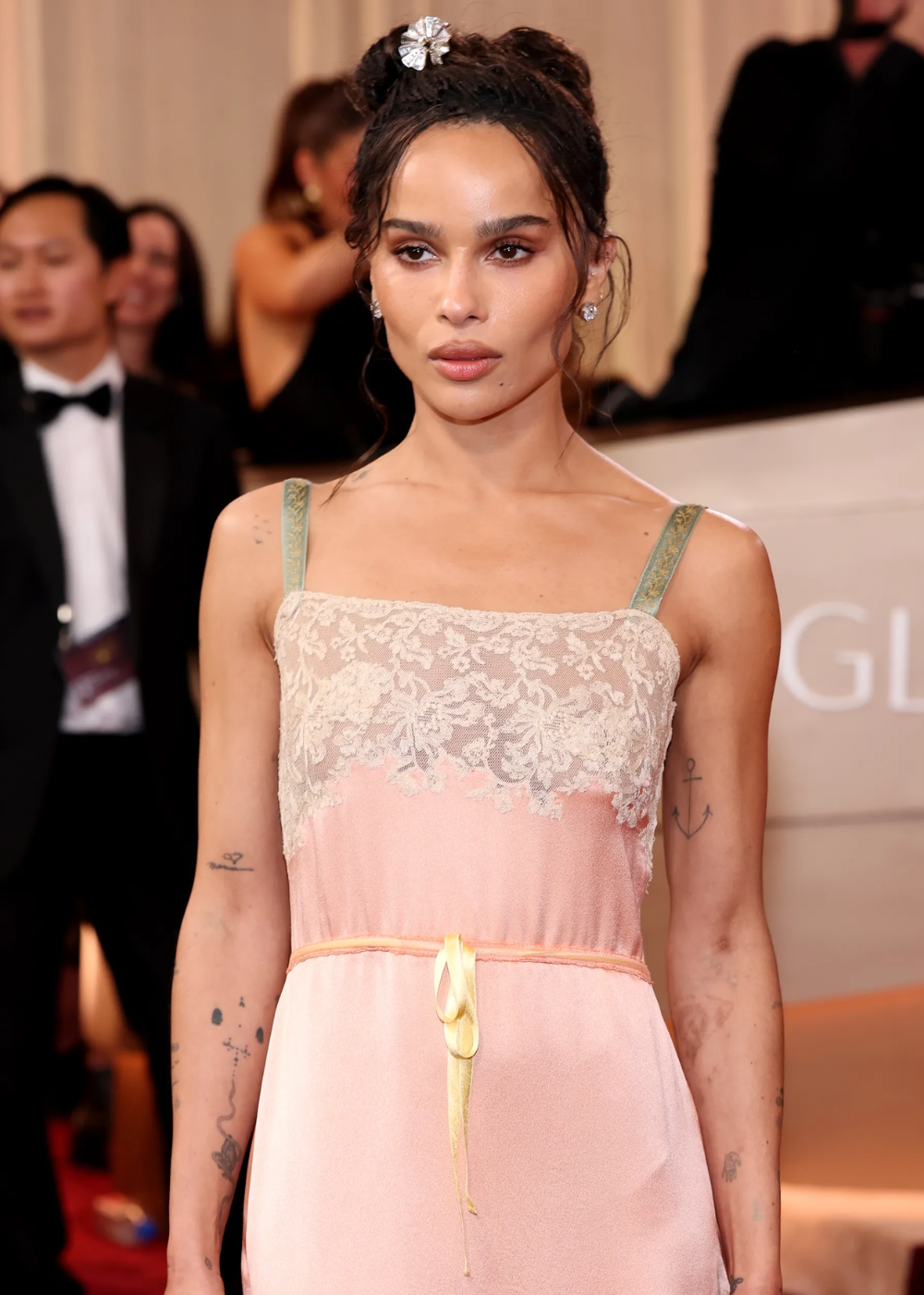 Modelo posa para o Globo de Ouro 2026 com vestido rosa claro em seda, renda branca no busto, alças verdes bordadas e laço.