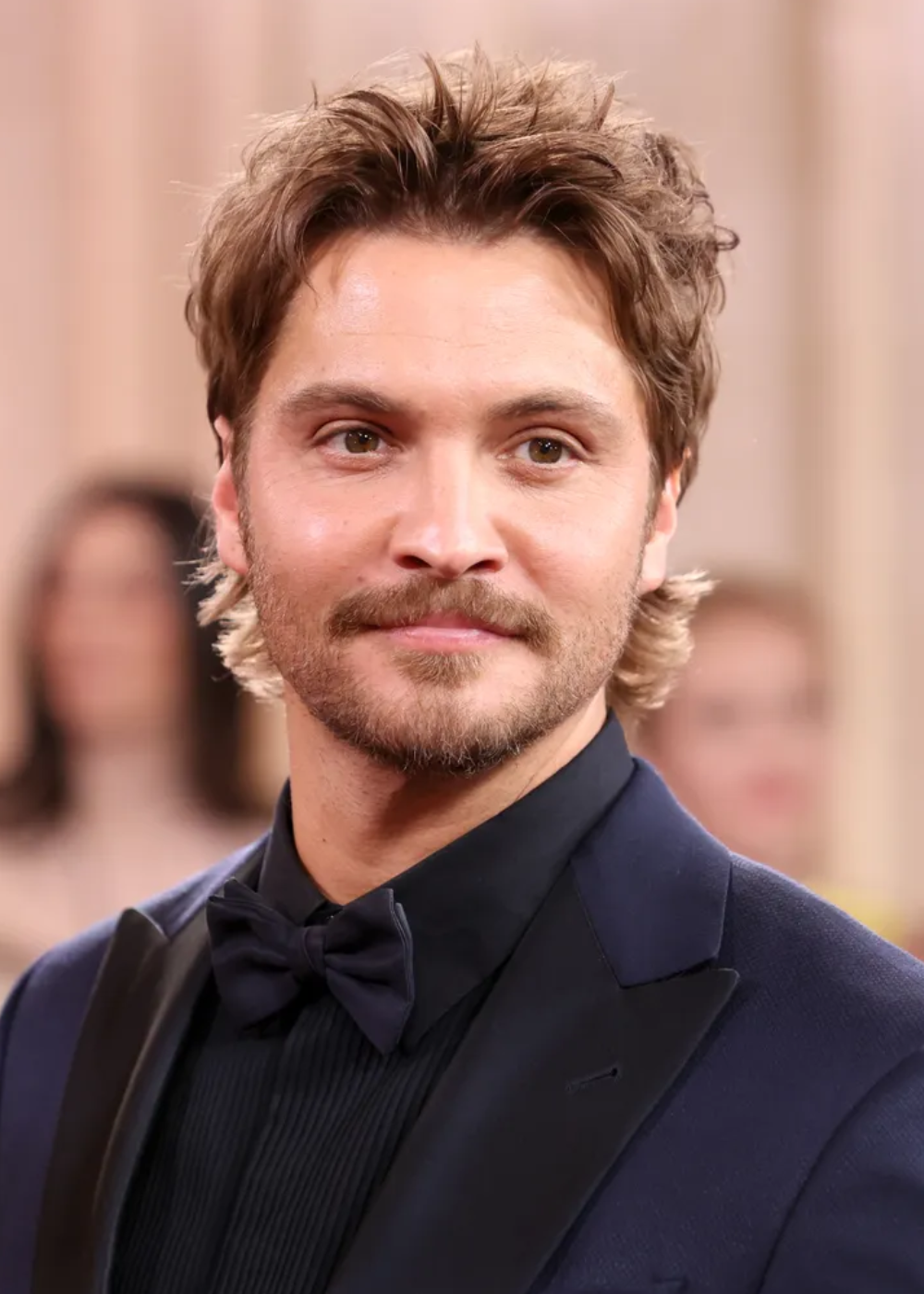 Ator posa com blazer azul marinho de corte clássico, camisa preta e gravata borboleta no clima elegante.