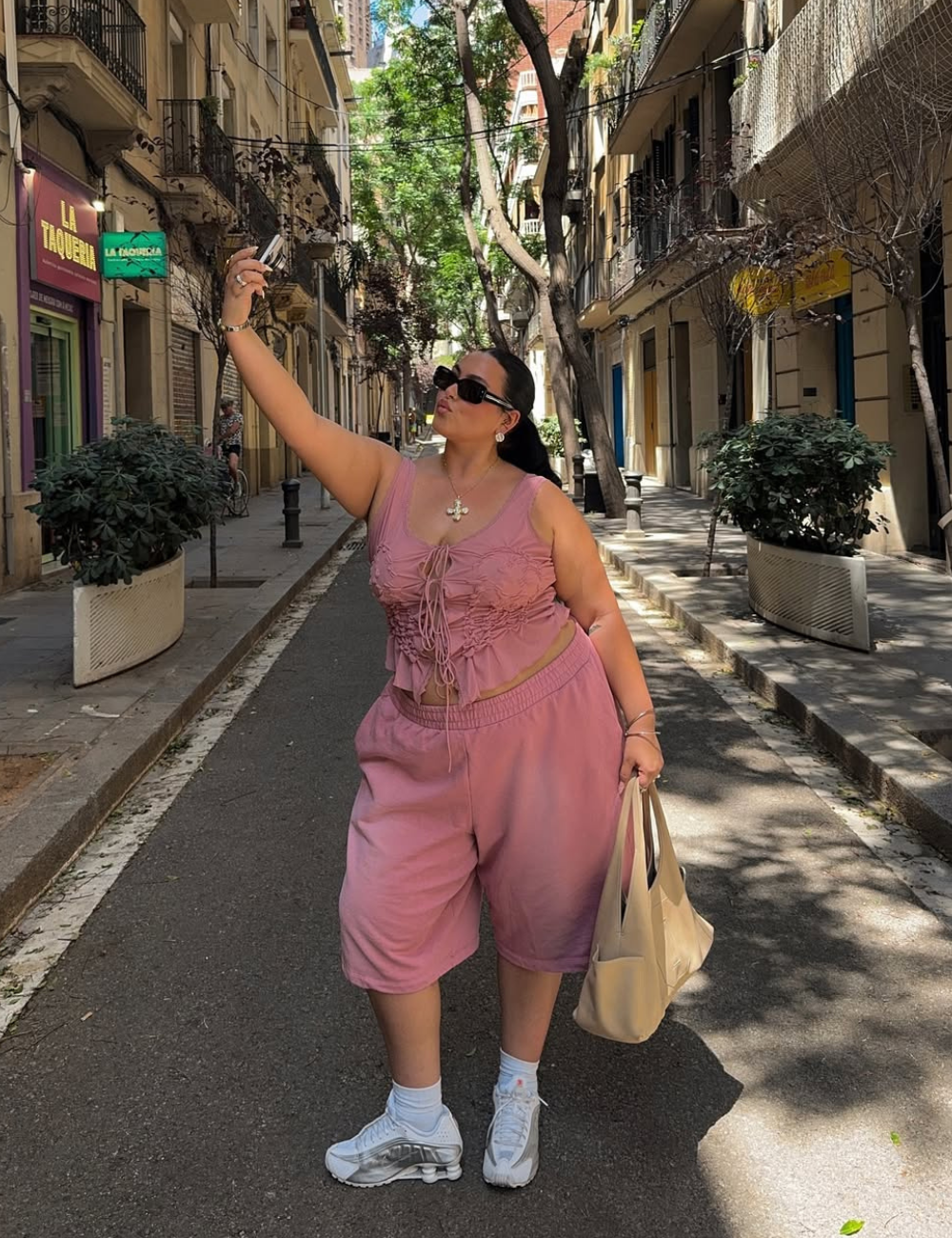 Mulher posa para selfie com tênis esportivo feminino branco, bermuda e top rosa em tecido leve, clima urbano e descontraído.