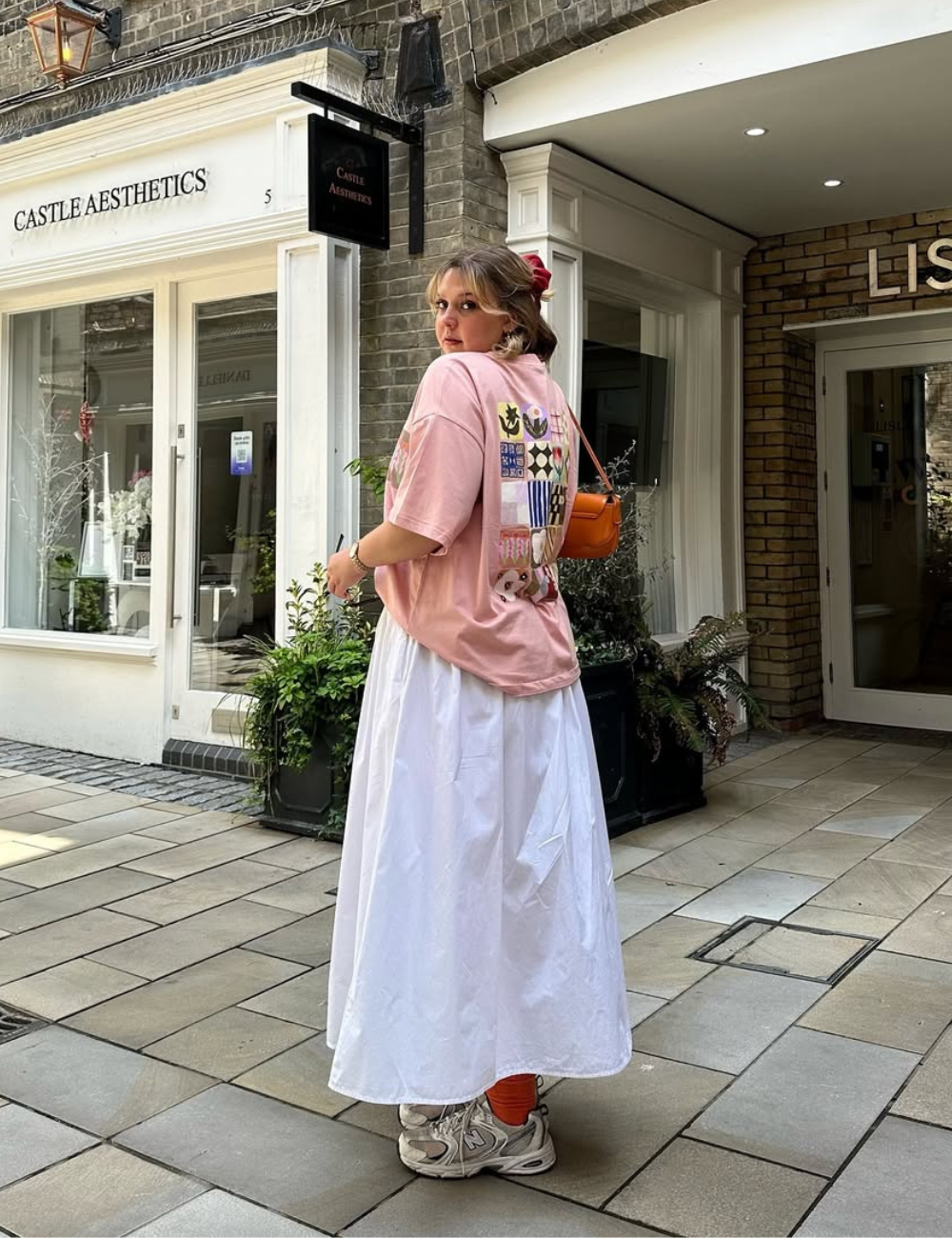 Modelo posa de costas em cenário urbano com tênis esportivo feminino branco, saia longa branca fluida e camiseta rosa.