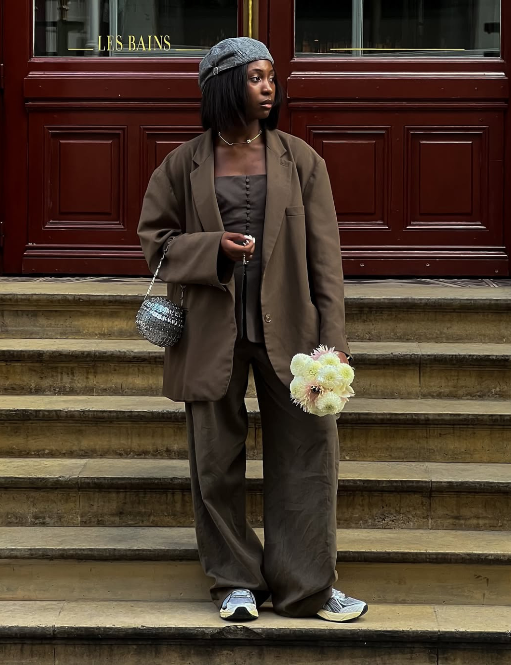 Modelo posa com blazer oversized e calça larga marrom, top tomara que caia, boina cinza e tênis esportivo feminino prata.