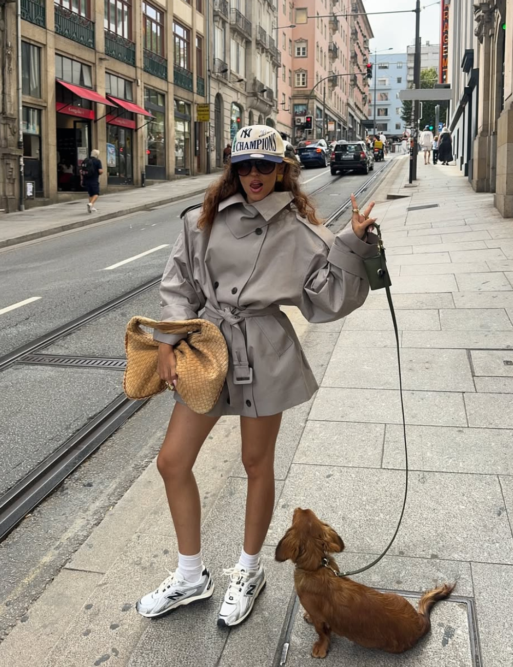 Modelo posa na rua com tênis esportivo feminino branco e cinza, meia branca, trench coat bege oversized e bolsa de palha.