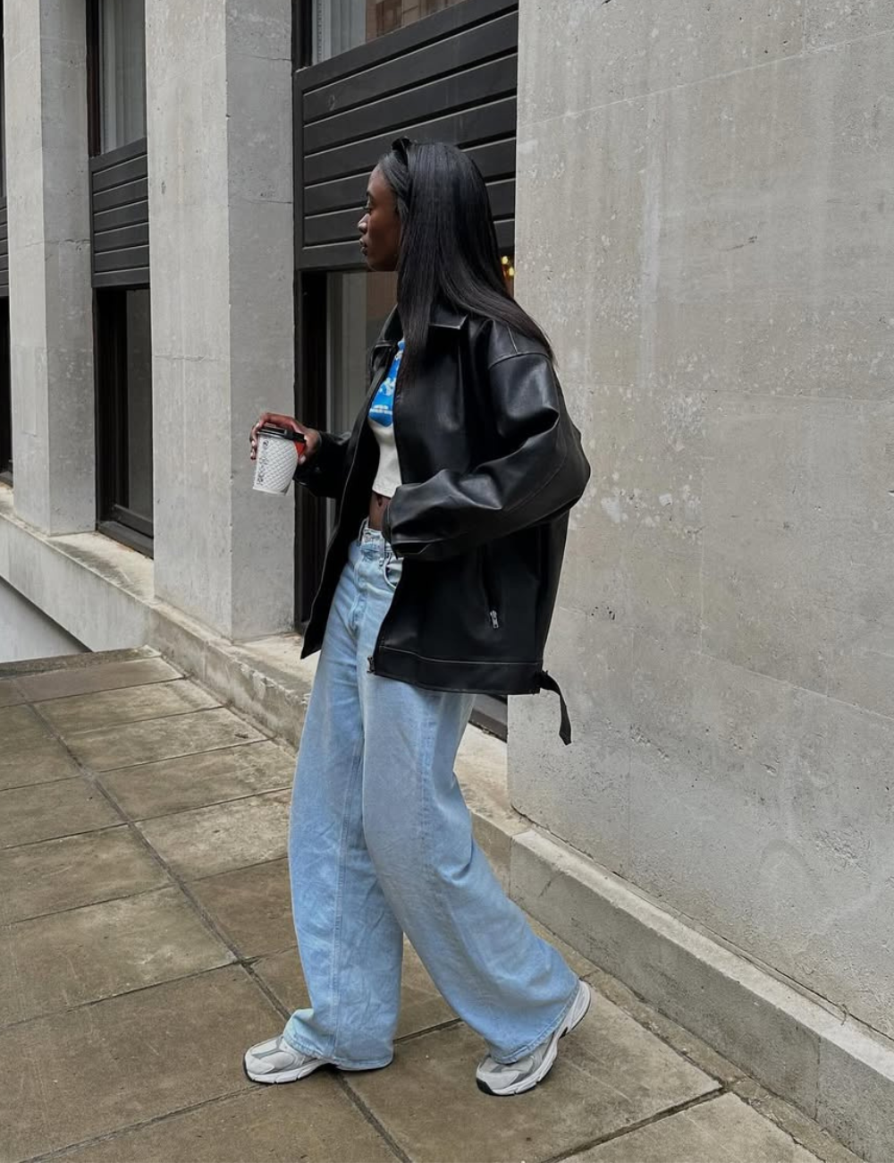 Mulher caminha segurando copo, veste jaqueta preta oversized de couro, cropped branco com azul, jeans claro largo e tênis.