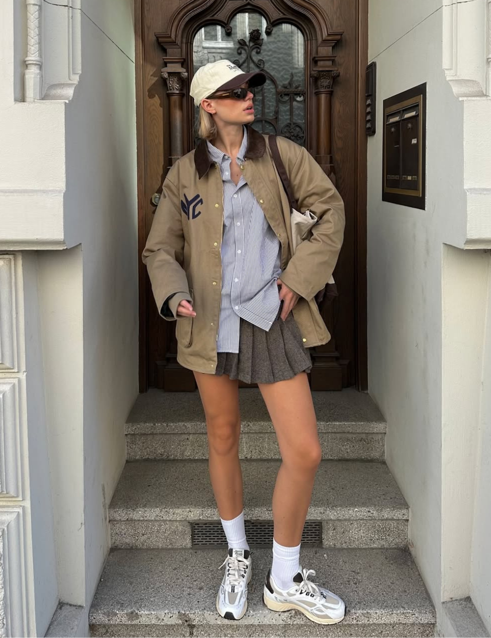 Modelo posa com tênis esportivo feminino branco com detalhes cinza e bege, combinando com meia branca e saia plissada marrom.