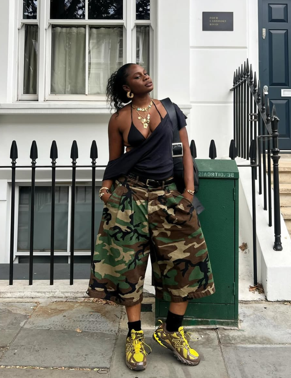 Modelo posa com atitude confiante usando tênis esportivo feminino amarelo e marrom, bermuda camuflada larga e blusa preta.