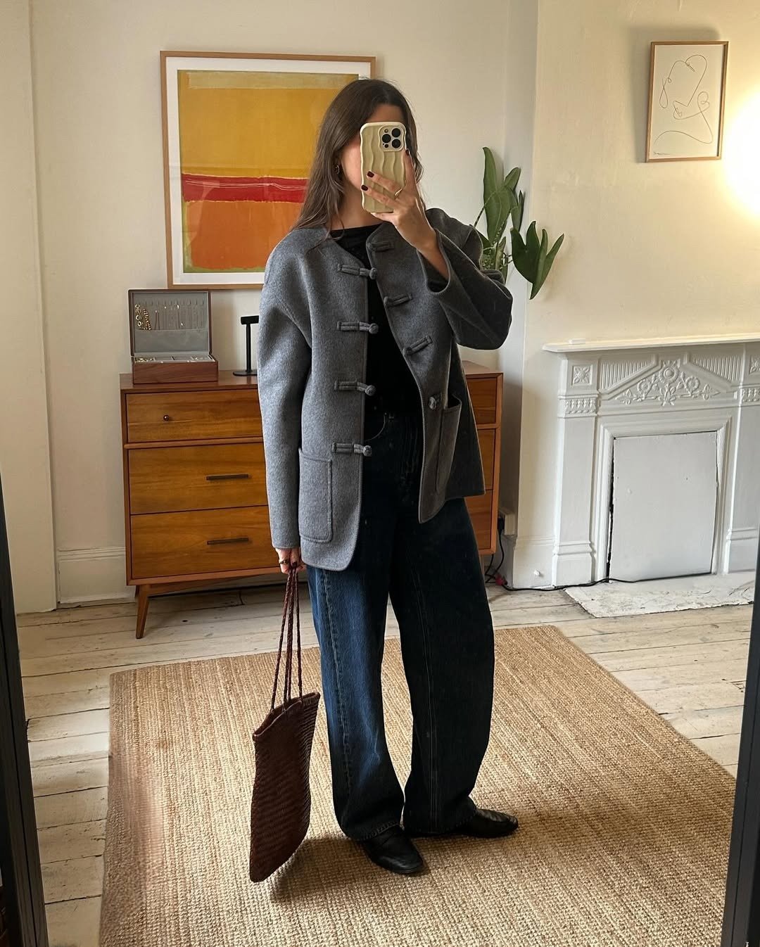 Modelo posa em ambiente aconchegante com casaco cinza de lã estruturado com nó chinês, jeans escuro e bolsa marrom trançada.