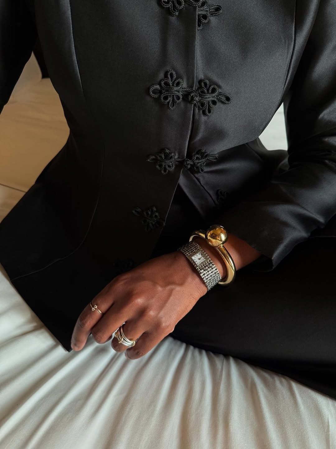 Blazer preto ajustado em tecido acetinado destaca o nó chinês em cordão fino, com pulseiras dourada e prata e anéis