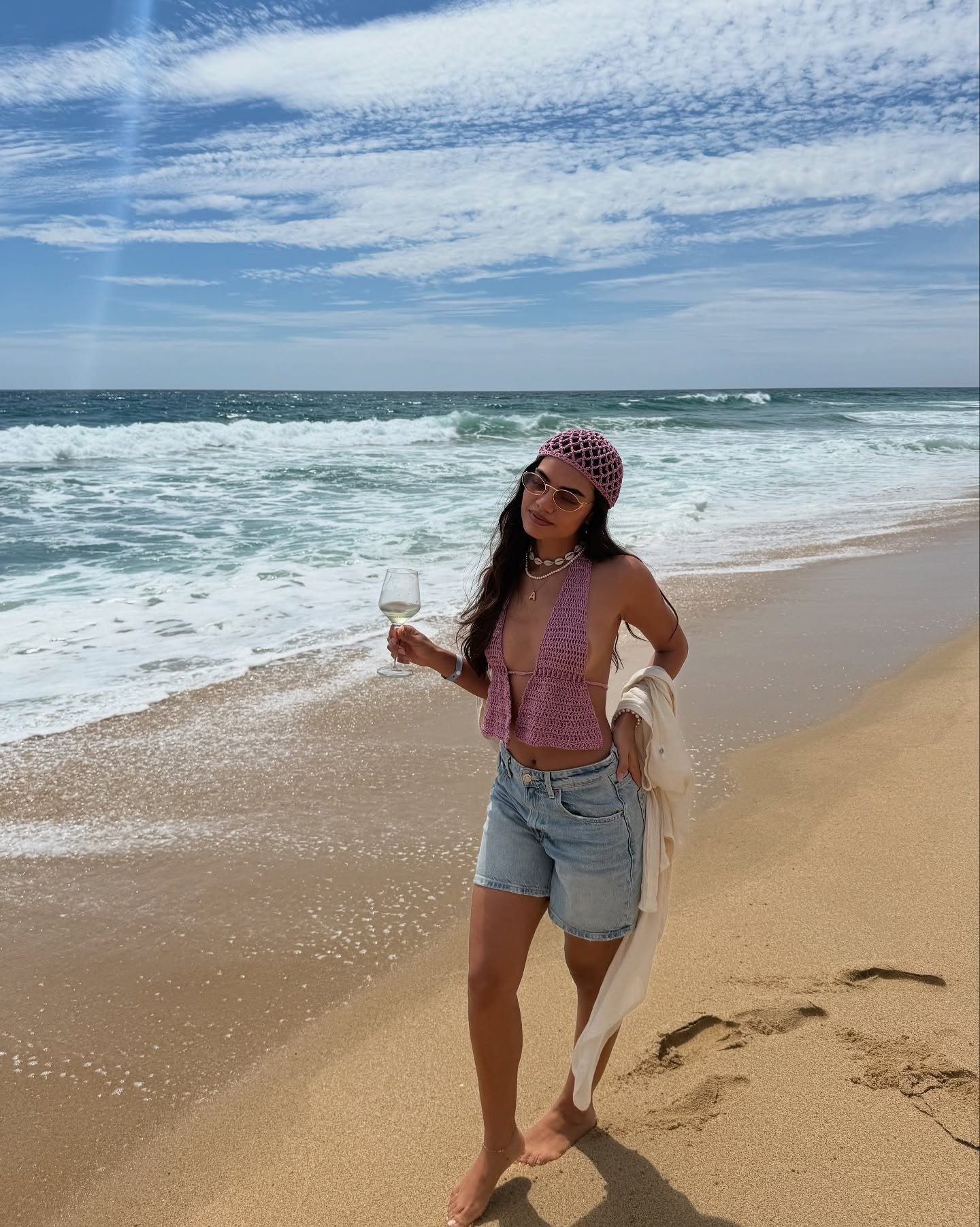 Modelo caminha na praia com top de crochê rosa em modelagem frente única, combinando com chapéu da mesma textura e shorts