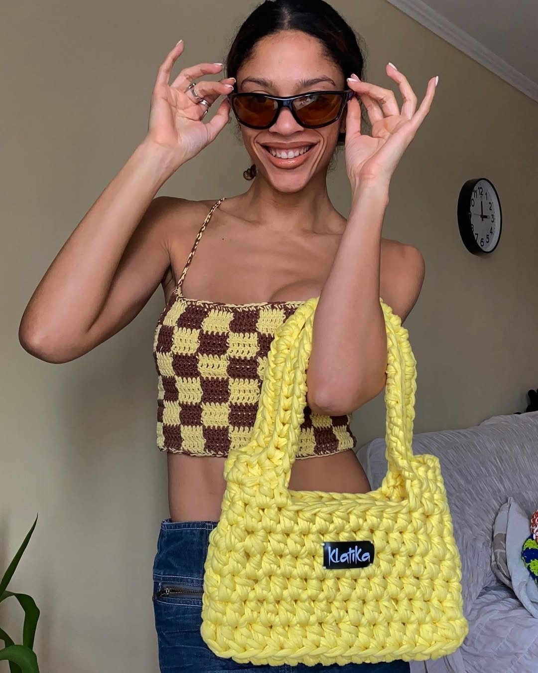 Modelo sorri segurando óculos escuros e bolsa amarela de crochê, vestindo top de crochê xadrez amarelo e marrom