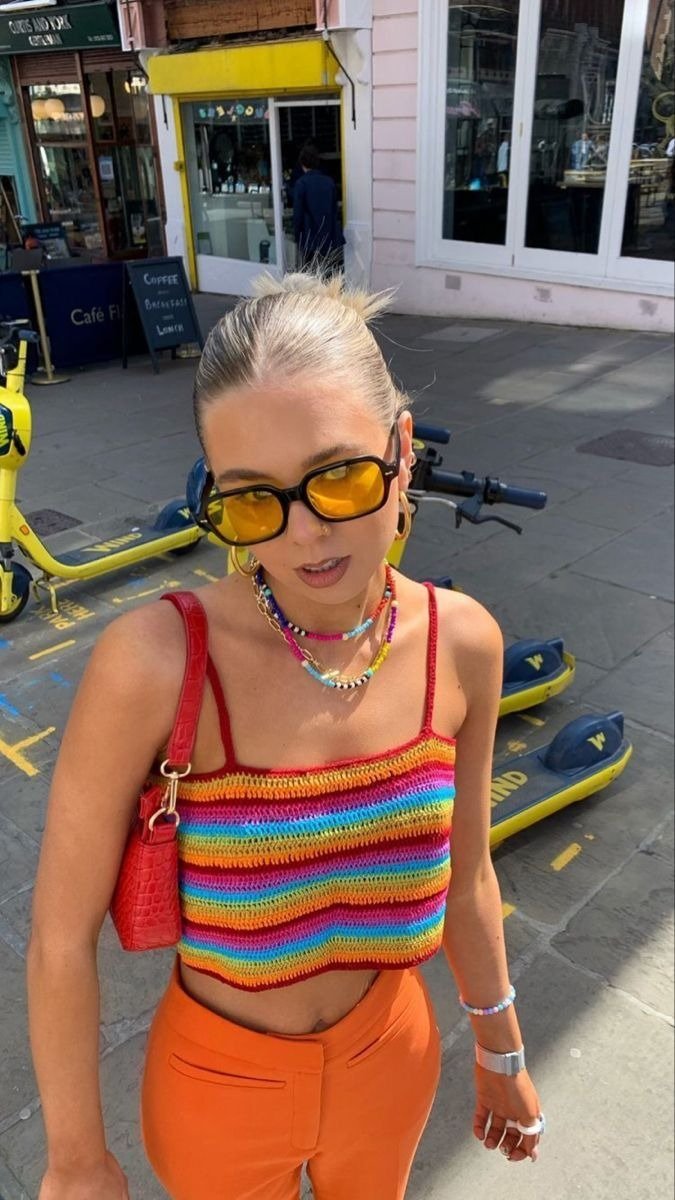 Modelo posa com top de crochê listrado em cores vibrantes, combinando com calça laranja e acessórios coloridos
