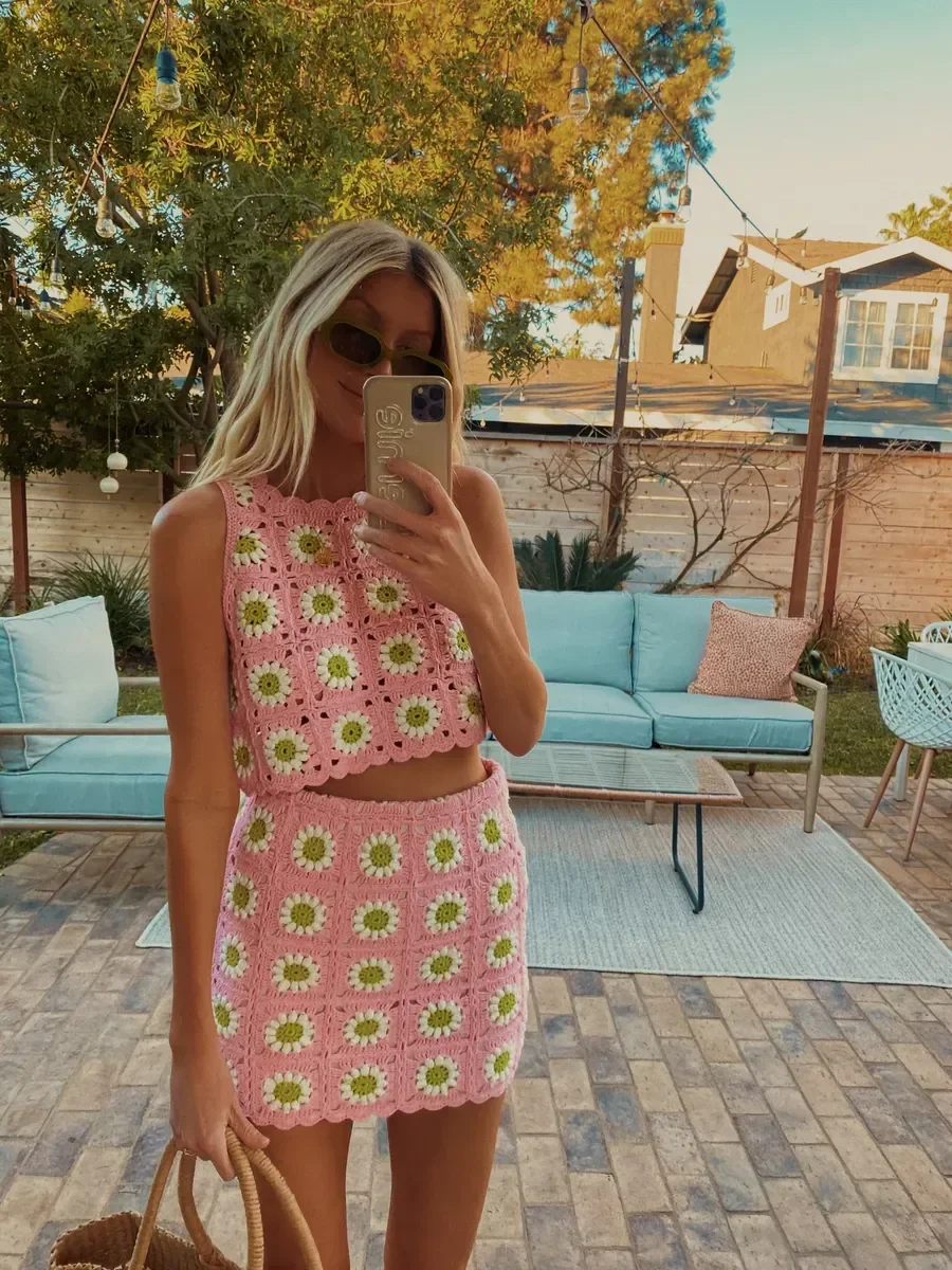 Modelo posa ao ar livre usando top de crochê rosa com flores brancas e verdes, combinando com saia curta e clima