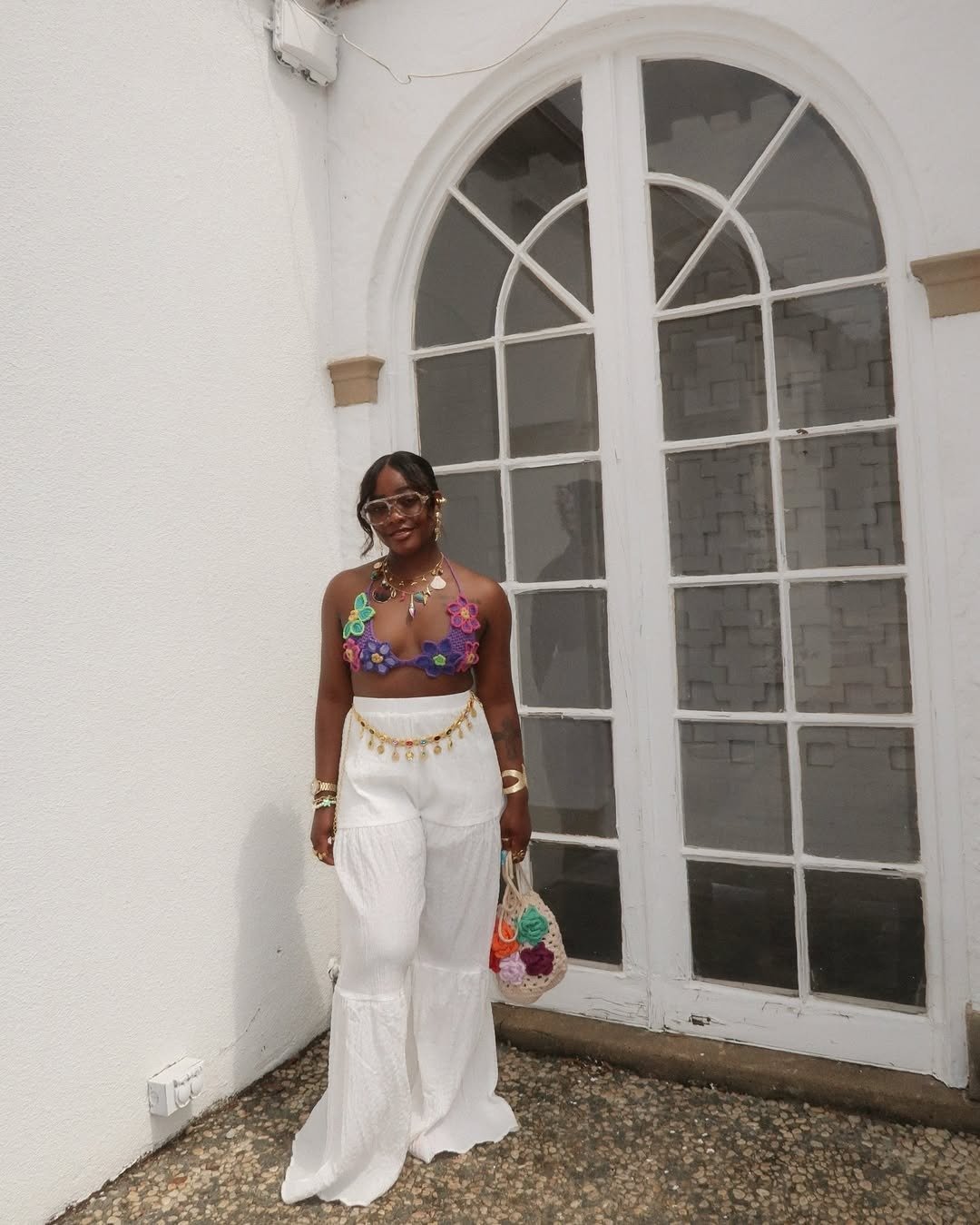 Modelo posa com top de crochê roxo decorado por flores coloridas, calça branca ampla e acessórios dourados, em clima leve e