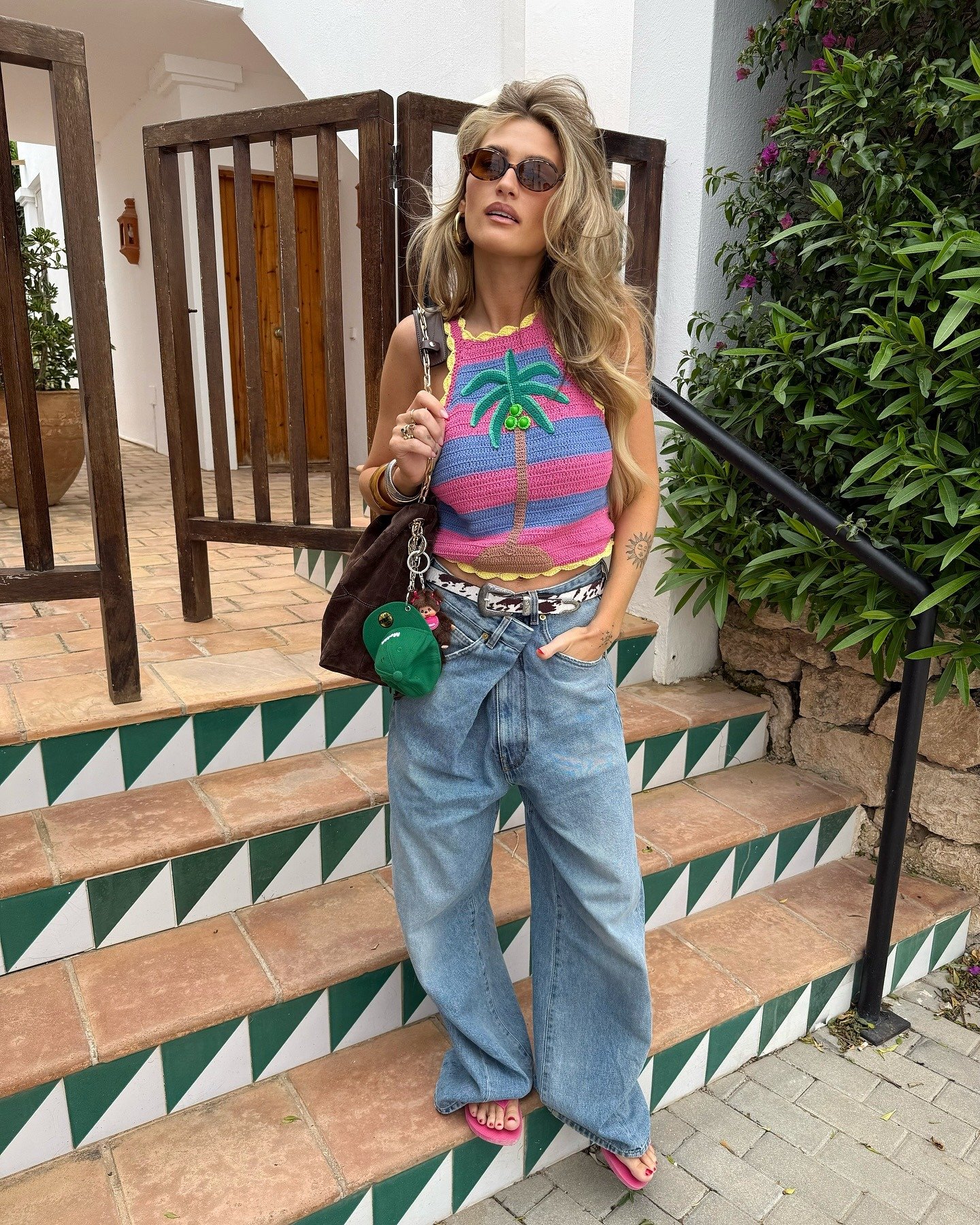 Modelo posa com top de crochê listrado rosa e azul, estampa de coqueiro, combinando calça jeans larga e acessórios casuais.