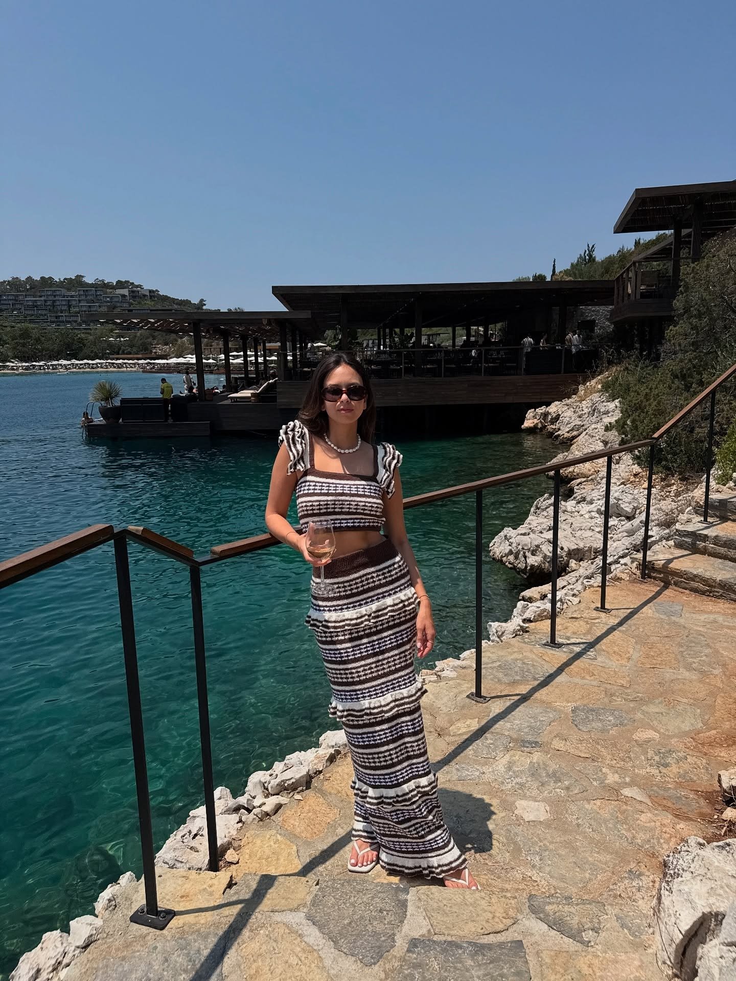 Modelo posa à beira-mar com top de crochê cropped listrado em tons neutros e saia longa combinando, clima elegante 