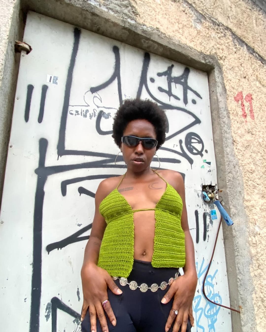 Modelo posa com top de crochê verde aberto na frente, combinando com calça preta e cinto de moedas prateadas