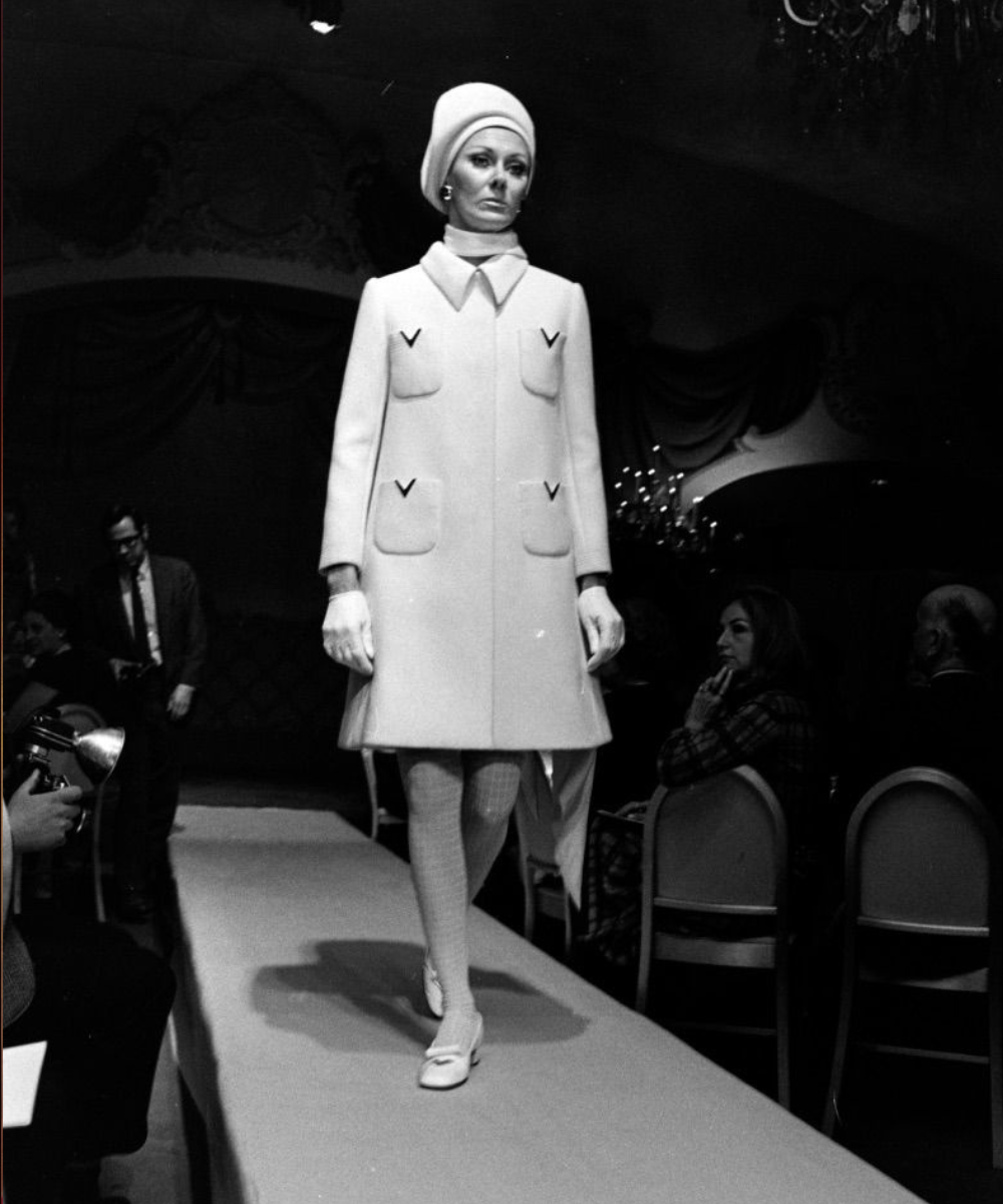 Modelo Valentino desfila com casaco branco de modelagem reta, bolsos frontais e detalhes em V, combinando boina e luvas, em