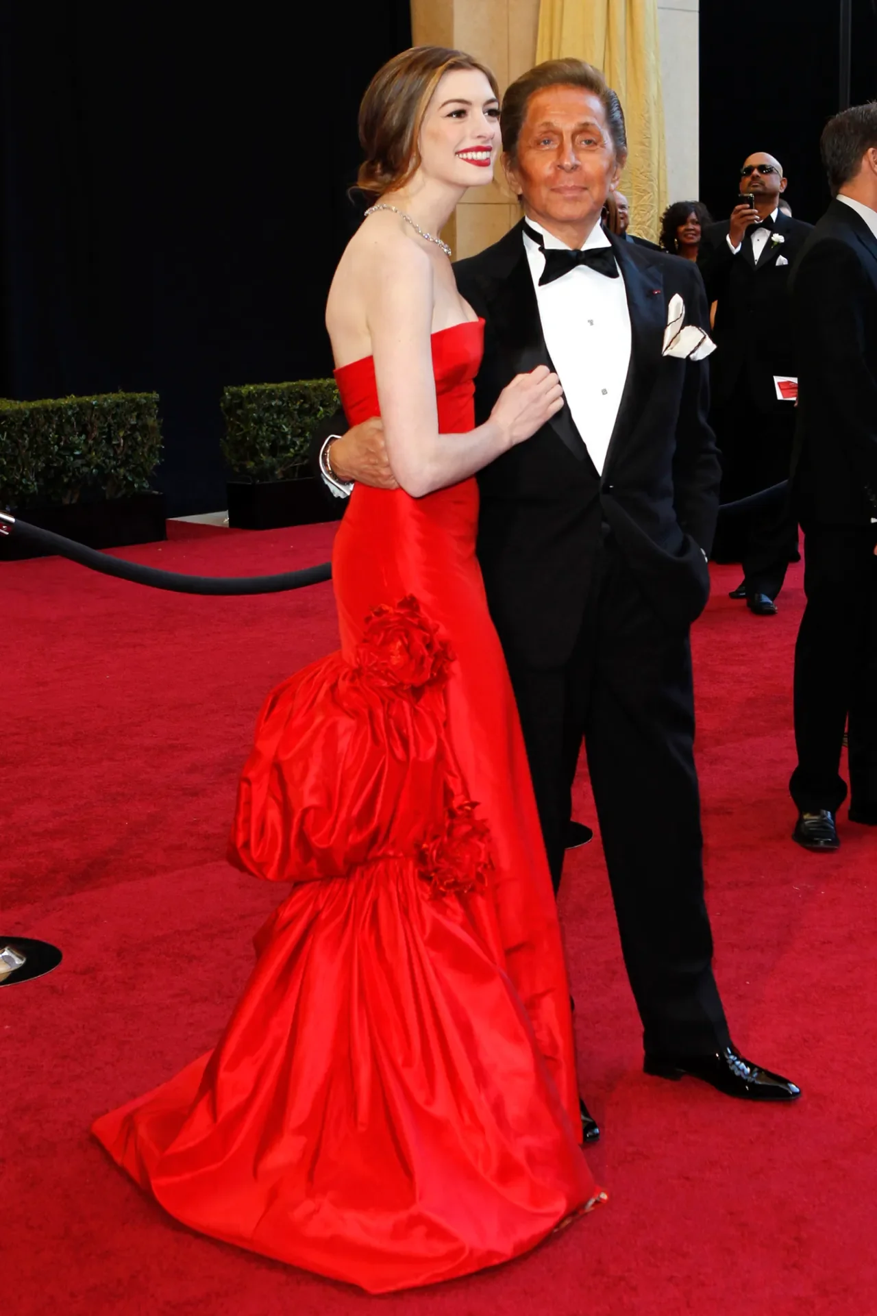 Modelo posa sorridente com vestido Valentino Garavani vermelho vibrante, tomara que caia, com drapeados e flores, ao lado de