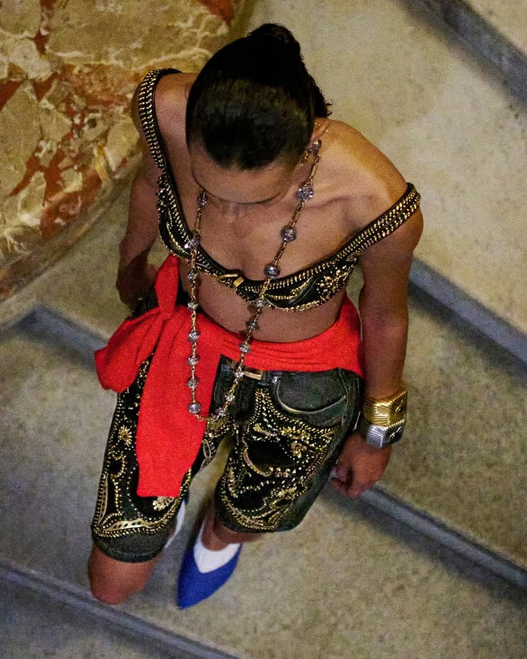 Modelo caminha em escada com top cropped e bermuda preta bordados em dourado, detalhes em vermelho vibrante e sapato azul,