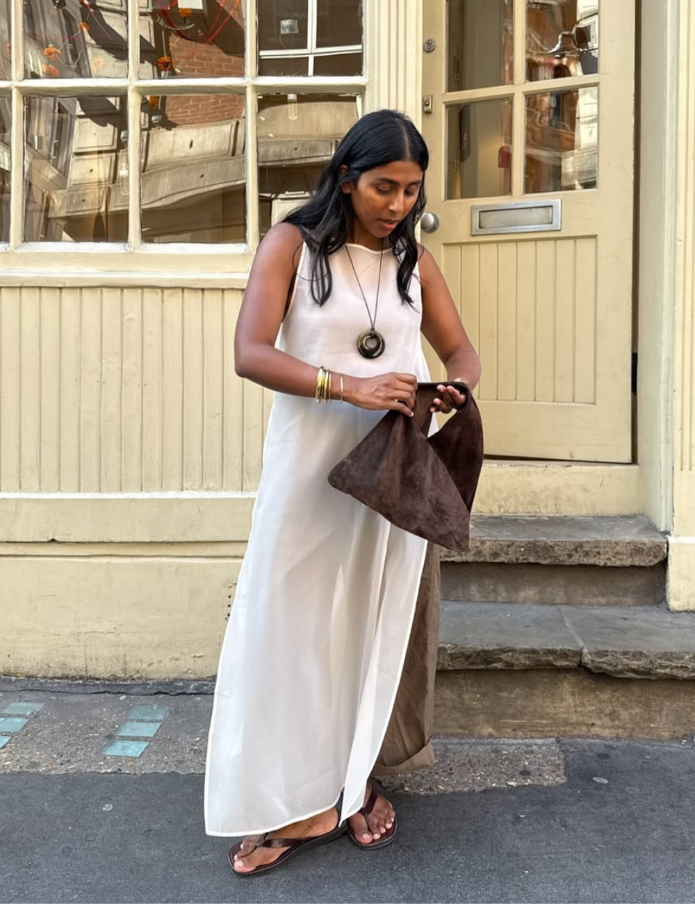 Modelo segura bolsa marrom em vestido longo reto branco de tecido leve, com acessórios dourados, em cenário urbano tranquilo.