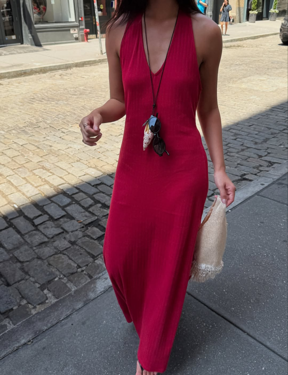 Modelo caminha com vestido longo reto vermelho em tecido leve e textura canelada, combinando colar com conchas e bolsa.