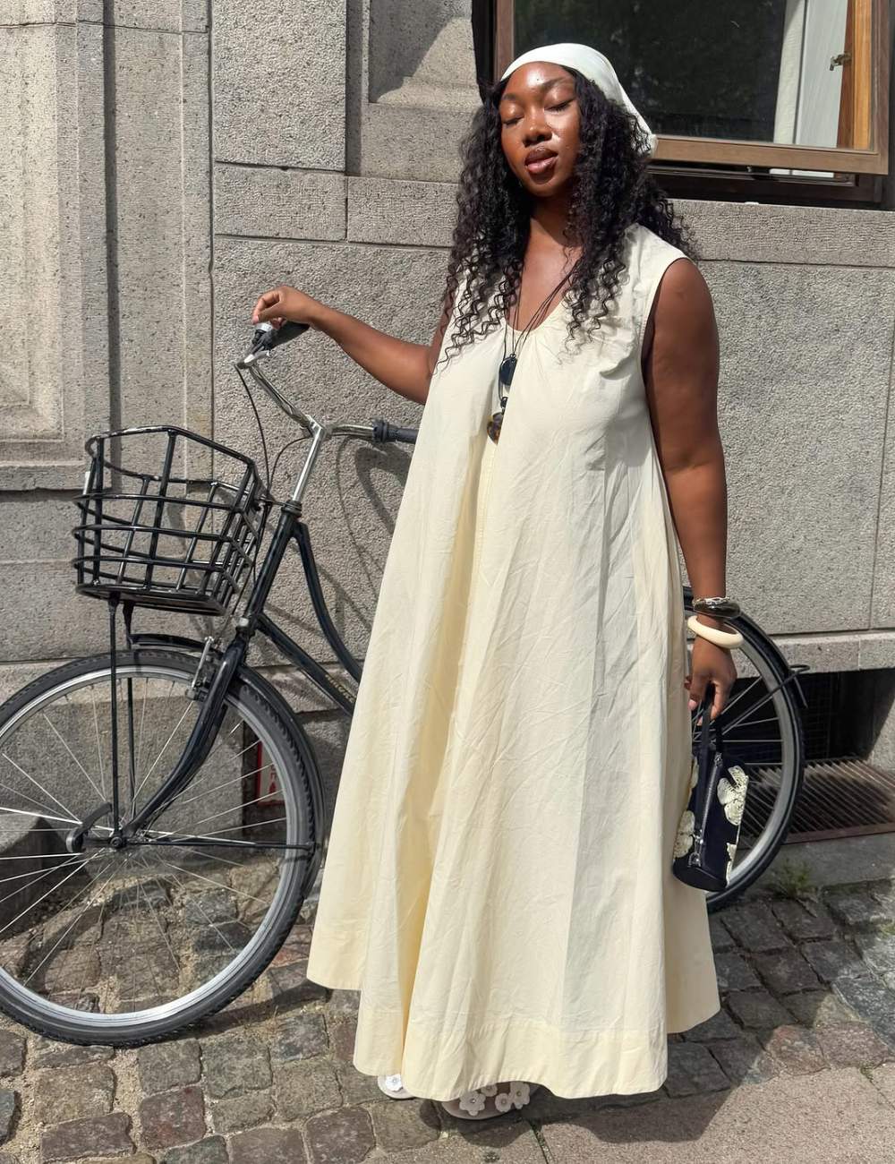 Modelo posa com vestido longo reto em tom creme, tecido leve e fluido, combinando acessórios neutros e bolsa preta.