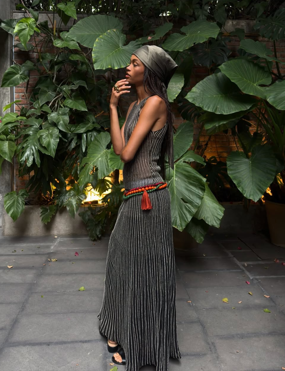 Modelo posa de perfil com vestido longo reto listrado em preto e branco, faixa colorida na cintura e lenço verde.