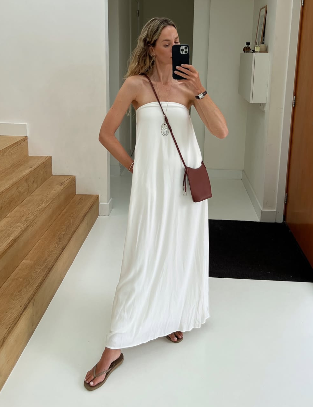 Modelo posa com vestido longo reto branco em tecido fluido, combinando bolsa marrom e sandálias rasteiras.