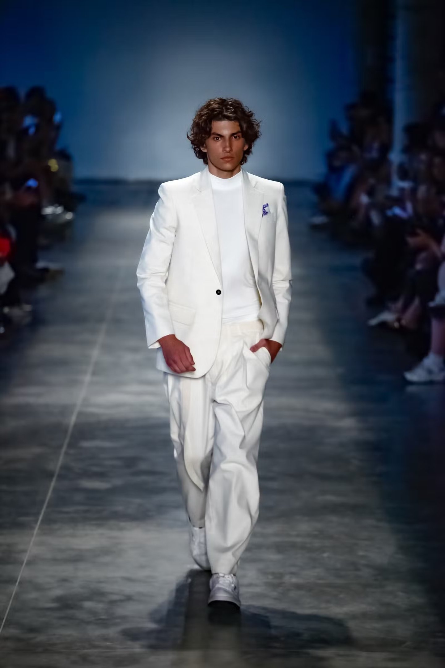 Homem caminha confiante com terno branco de modelagem ampla, camiseta gola alta e tênis, cenário sóbrio realça elegância