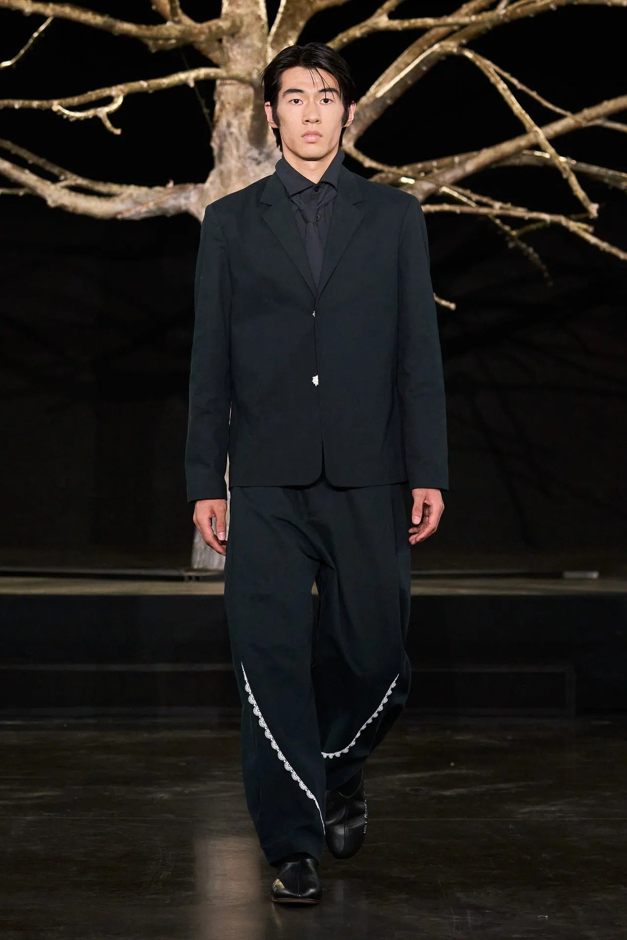 Wagner Moura no Oscar veste terno preto de modelagem ampla com detalhes brancos nas calças, postura séria e clima sóbrio.