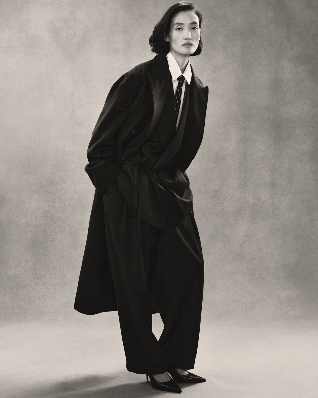 Modelo posa com postura confiante usando sobretudo e alfaiataria oversized em tons escuros, refletindo a indústria da moda