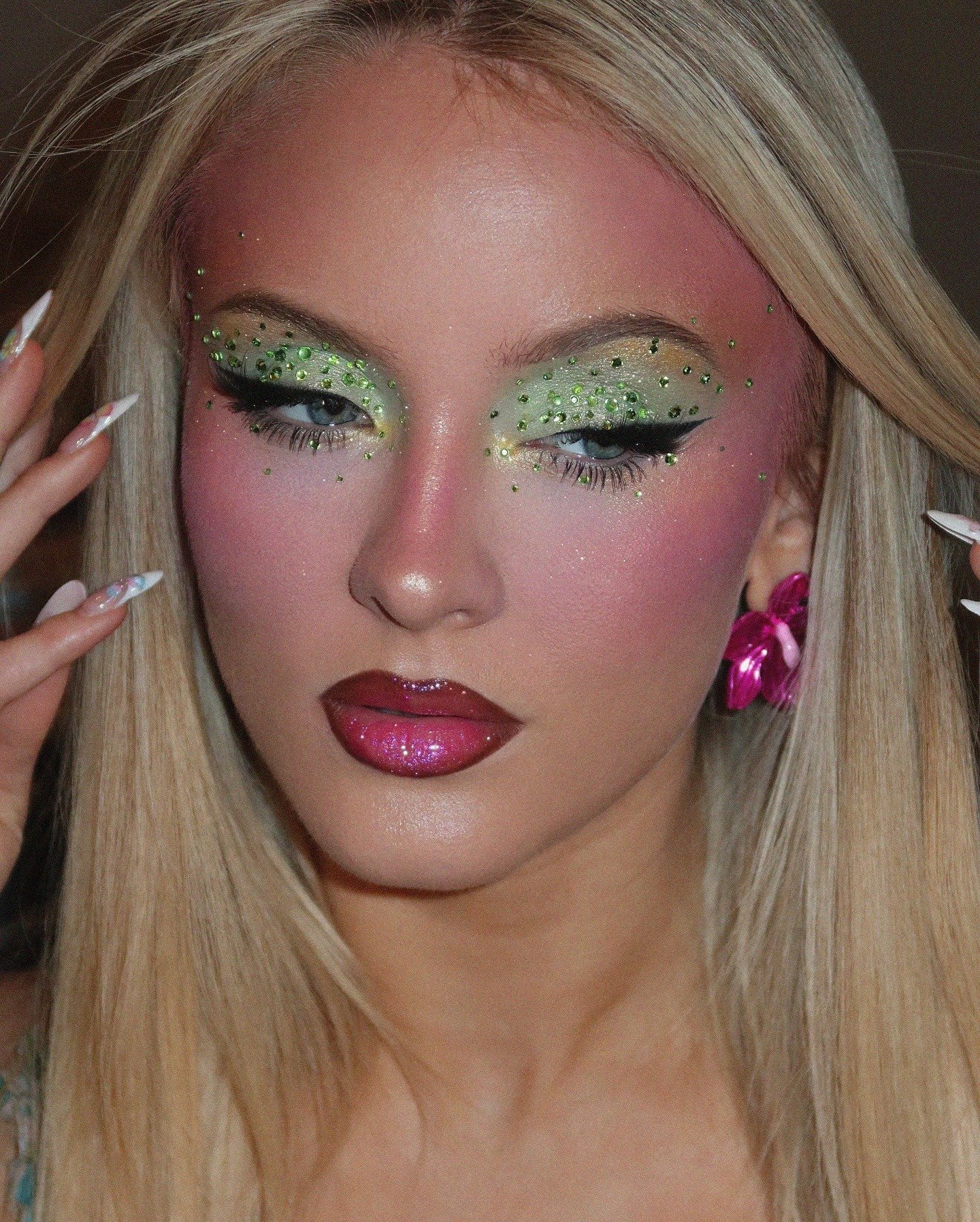 Zara Larsson exibe maquiagem artística com sombra verde e cristais, batom rosa vibrante e brincos florais, em pose delicada