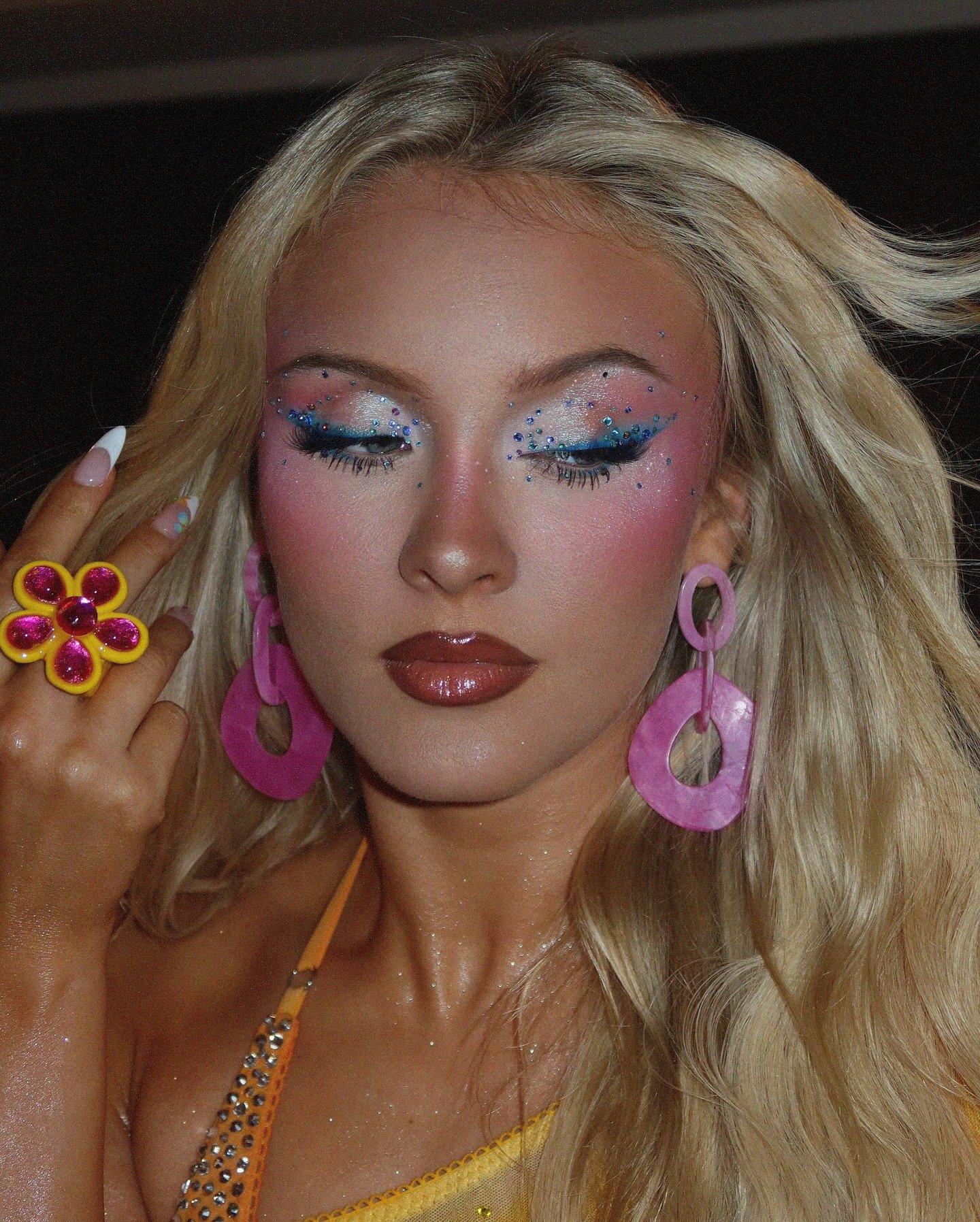 Zara Larsson exibe maquiagem vibrante com glitter azul, brincos grandes rosa, anel florido amarelo e top amarelo com strass