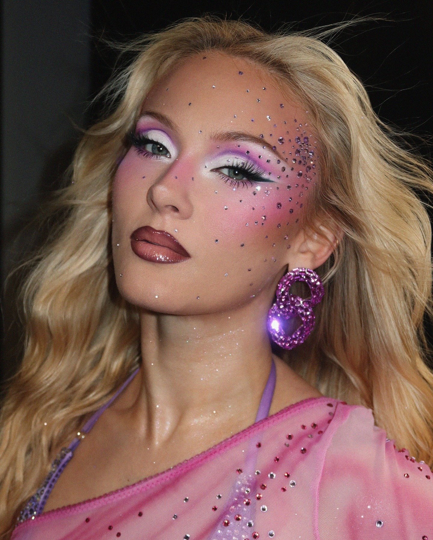 Zara Larsson exibe maquiagem artística em tons de lilás e rosa com cristais, brincos grandes e look rosa translúcido