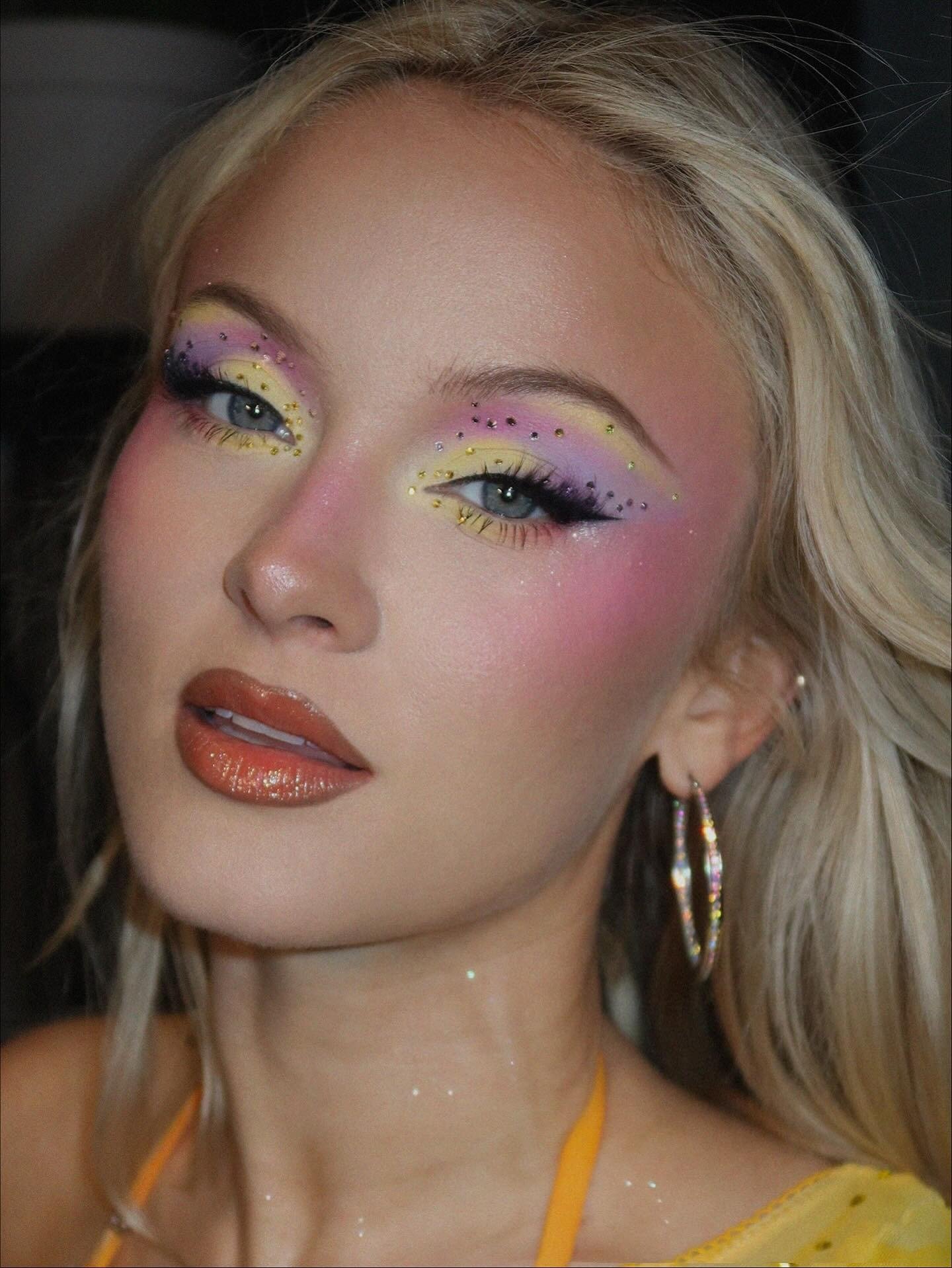 Zara Larsson exibe maquiagem artística em tons de amarelo e lilás com cristais, batom marrom e brincos de argola brilhantes