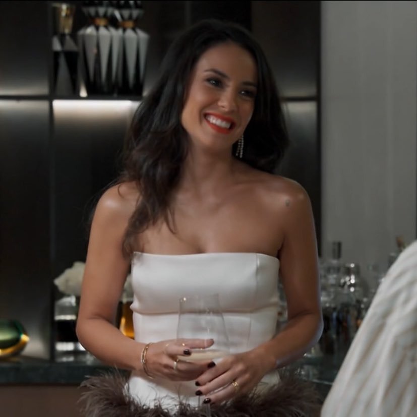 Zenilda sorri confiante, vestindo vestido branco tomara que caia com saia de plumas marrons, em ambiente sofisticado