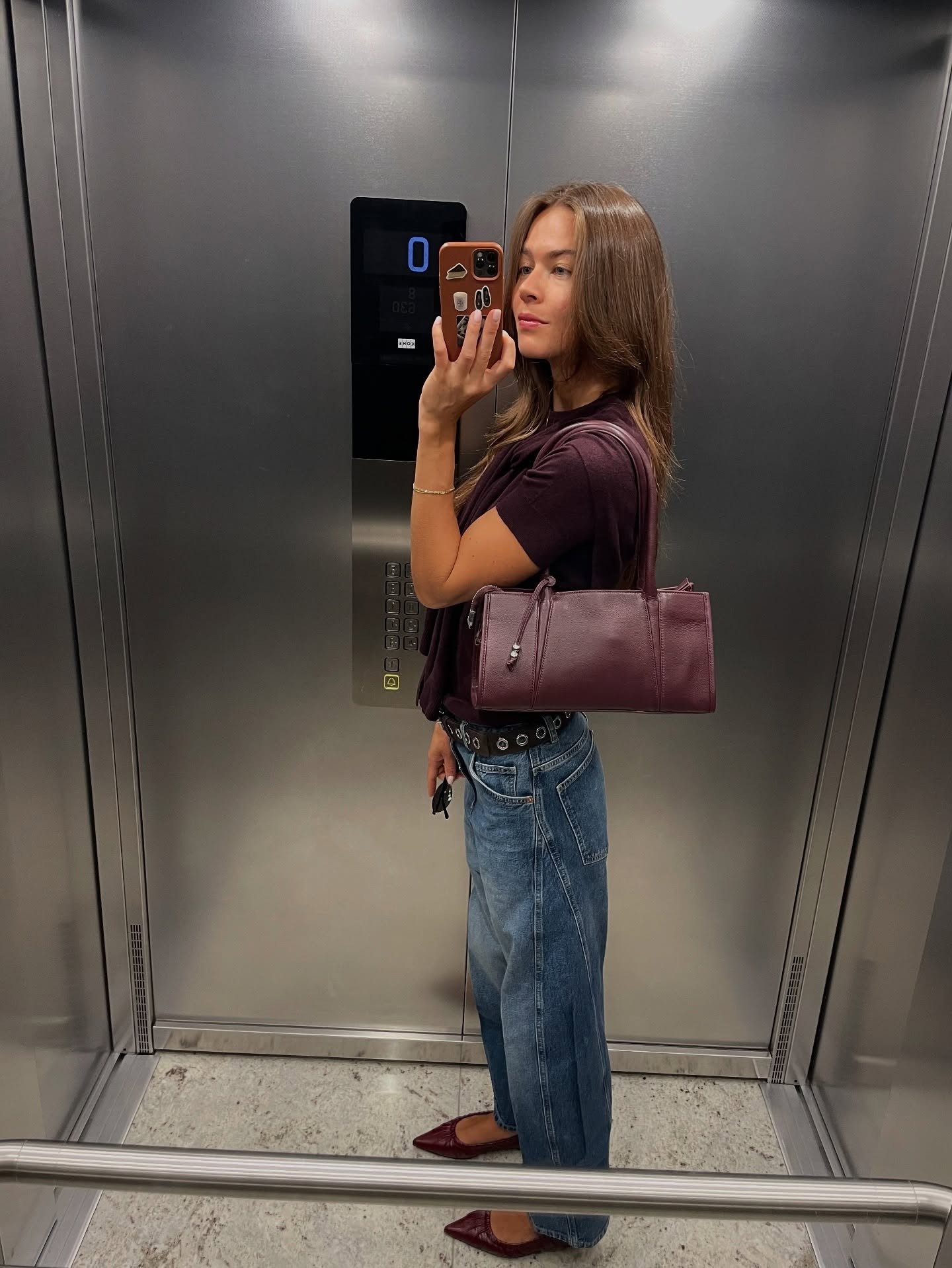 Modelo posa no espelho do elevador com looks bonitos em blusa vinho de malha, jeans amplo, bolsa e sapatilhas combinando.