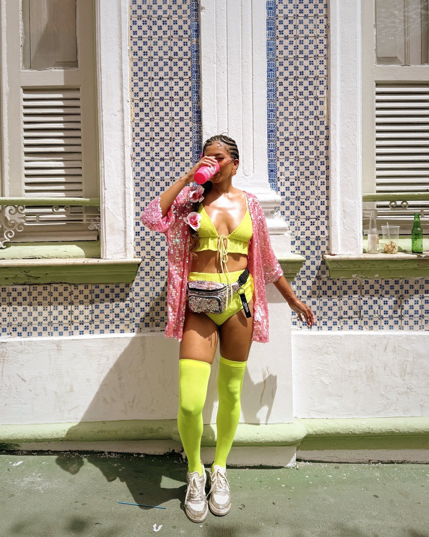 Modelo bebe de copo rosa usando biquíni amarelo neon com meia longa da mesma cor, kimono rosa de paetês e pochete brilhante