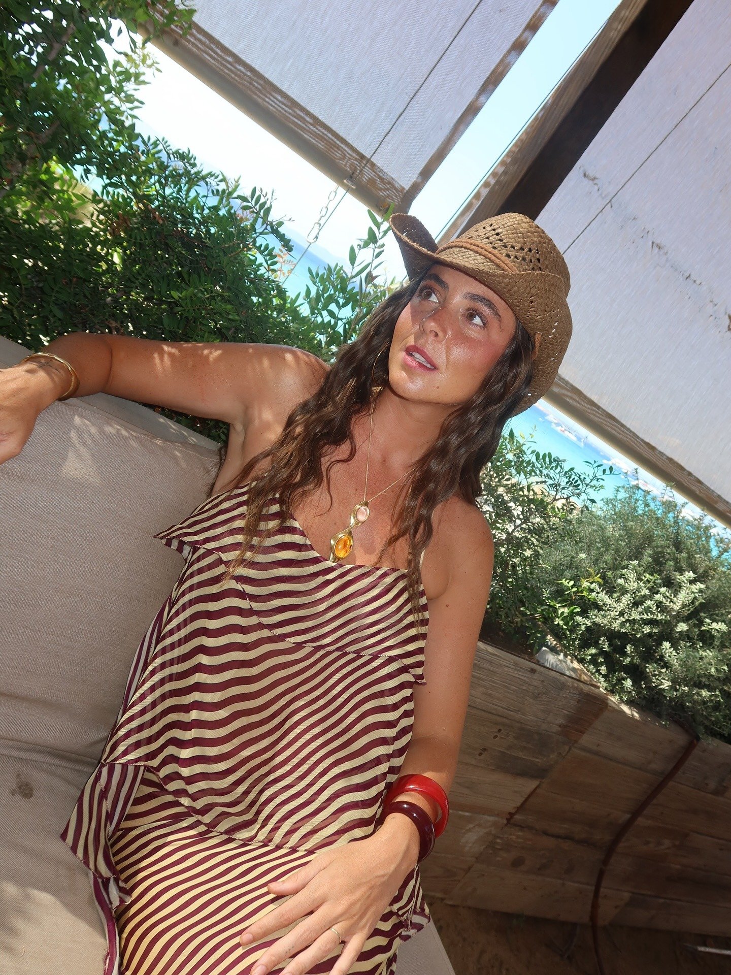 Modelo posa relaxada com vestido listrado vinho e bege, chapéu de palha e acessórios tendência de 2026 em resina vermelha