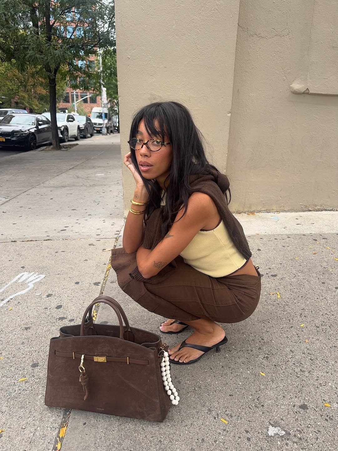 Modelo agachada com tricô leve marrom sobre os ombros, regata bege e calça marrom, em clima casual urbano e sofisticado.